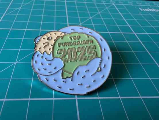 An enamel pin that says Top Fundraiser 2025 on it and there is an otter cuddling the letters.
