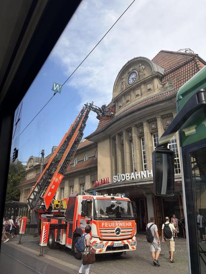 The image shows a fire truck with an extended ladder positioned in front of a historic building labeled "SÜDBAHNH." People are walking nearby, and the scene is set under a partly cloudy sky with a visible clock on the building.