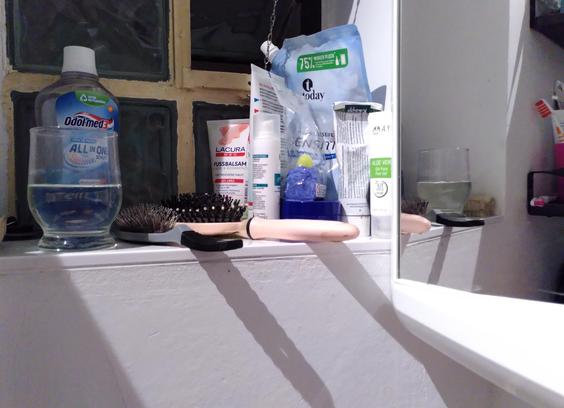 Fenstersims in einem Badezimmer, unter anderem mit Haarbüsten, diversen Tuben und einem Glas Wasser, daneben das Ganze im Badezimmerspiegel. Dort sieht das Wasser gelblich aus, weil eine beigefarbene Nagelbürste dahinter liegt. Im Original wirkt das Wasser blau, weil es in dieser Richtung vor einer Flasche Mundwasser steht.
