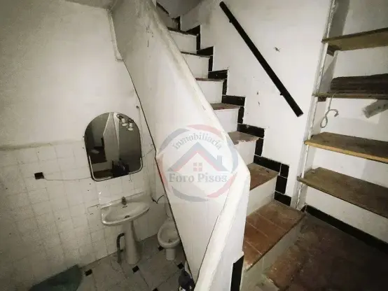 Foto de una habitación con una escalera que sube haciendo esquina. Se puede ver una pila con su espejo y un retrete en el hueco que queda debajo.