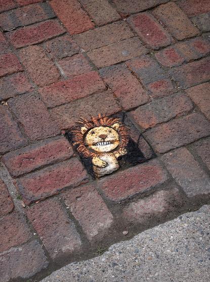sidewalk chalk drawing of a lion hand puppet emerging from an anamorphic hole in a brick walkway, showing her teeth