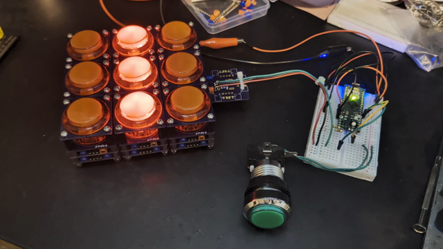 A 3x3 grid of illuminated arcade buttons, each a separate little tower of 4 PCBs only slightly wider than the button itself. A hand is pushing another button which is connected to an Arduino on a breadboard, which in turn is connected to the grid. The middle column of the grid is lit up, but with every pair of clicks from the button being pushed by hand, the middle column and the middle row alternate being lit up.