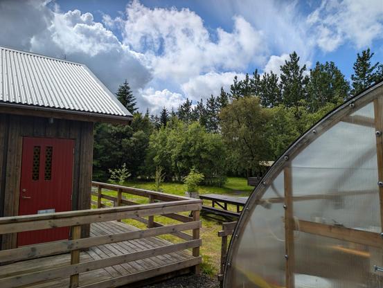 Cabin to the left, greenhouse to the right, green forest in the middle, under unsettled blue and cloudy sky.