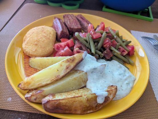 Ein runder gelber Teller, auf dem unteren Viertel Kartoffelspalten aus dem Airfryer, links oben das Cordon Bleu & 3 Steakstreifen, rechts oben der Tomaten-Bohnen-Salat und rechts unten der Skyr-Dip