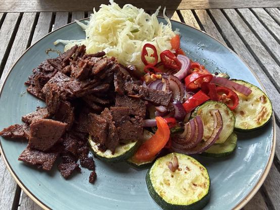 Ein Teller mit angebratenen Fleischstreifen, gegrillter Zucchini, Sauerkraut sowie roten Zwiebeln, Paprika und Chili auf einem Holztisch.