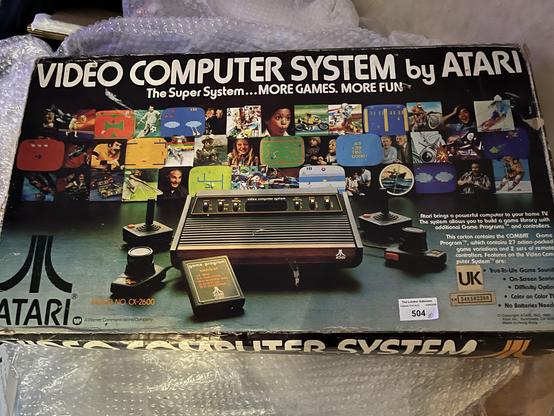An Atari video computer system box displays the slogan "The Super System... MORE GAMES. MORE FUN." The box features a vintage design with images of various games and players enjoying the console. The model number CX-2600 is visible