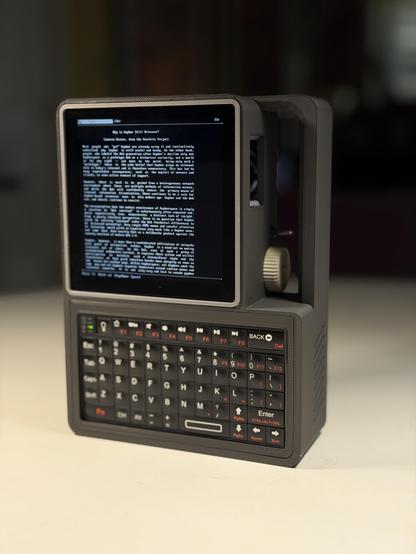 A small handheld computer in dark grey plastic standing up on a white table. It has a square LCD showing text from a Gopher site, a small keyboard at the bottom, and a handle on the top right.