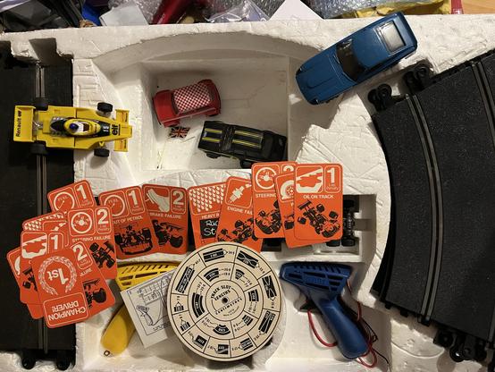 A collection of Scaletrix cars, including a yellow racing car, red mini with a checkerboard pattern, and a blue car. There are several orange cards labeled with various racing-related issues, a circular speedometer that looks like it calculates scale speed