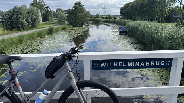 bicycle leaning against a tiny wooden bridge labeled "Wilhelminabrug (1908)" with a very pleasant dutch countryside in the background