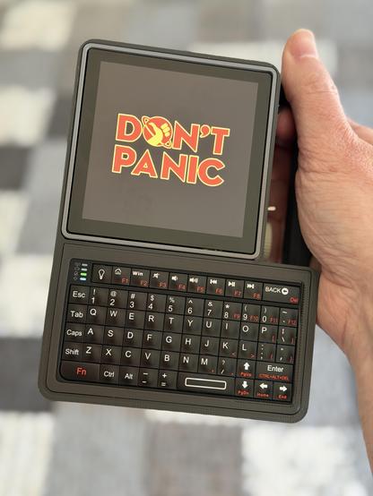 A hand holding a small computer by the handle with small keyboard at the bottom and display at the top left with the words "don't panic" in large friendly letters.