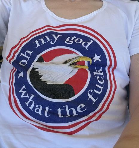 the torso of a person wearing a tshirt which mostly fills the whole frame. the shirt is white, with two thin concentric red circles bordering a wider blue circle. in the center is a drawing of a bald eagle with an extremely distressed expression on its face, its eyes wide and beak open. in the blue circle around the eagle, white letters read "oh my god what the fuck".