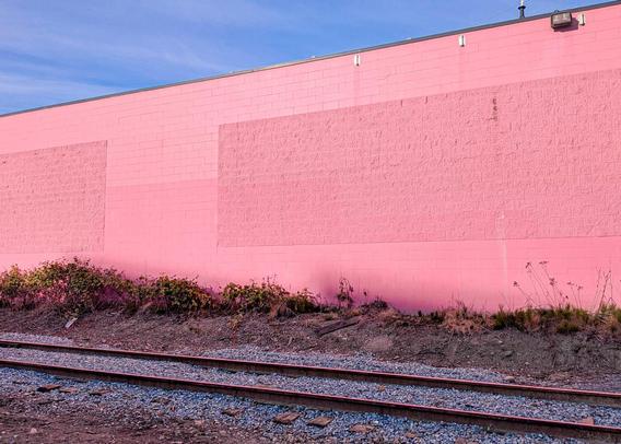 A large expanse of concrete-brick wall with an interesting pattern, painted an intense pink.  Above, blue sky, a few clouds. In teh foreground, bare industrial land traversed by a railway track which is reflecting the wall's pink.