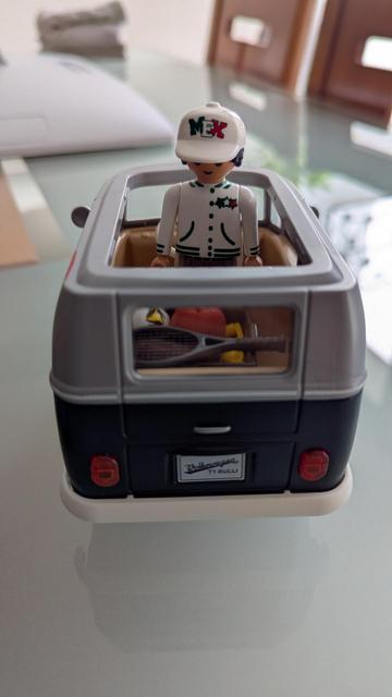 The VW bus from behind without the roof showing the interior. Playmobil dude is standing on the front seat. He's wearing a white hat with the word MEX on it and a white coat. On the back of the van thereis a box with a football, basketball and a tennis racquet