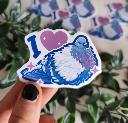 hand holding up a sticker of a resting pigeon titled "i <3 (pigeons)"