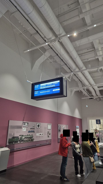 A DB LCD passenger information display hanging from the ceiling in an exhibition hall in a museum with people below. It shows show times for the model railway