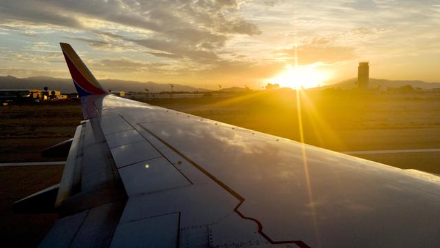 A Southwest Airlines aircraft wing extends across the frame during sunrise at Tucson airport, with the sun low on the horizon casting golden light across the tarmac, control tower, and distant mountains in the background.