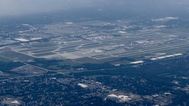 Aerial view of John Glenn Columbus International Airport with multiple parallel runways and taxiways, surrounded by forested areas and residential neighborhoods. The airport infrastructure is clearly visible with its distinctive runway layout, while wooded areas and scattered buildings occupy the surrounding landscape under hazy atmospheric conditions.
