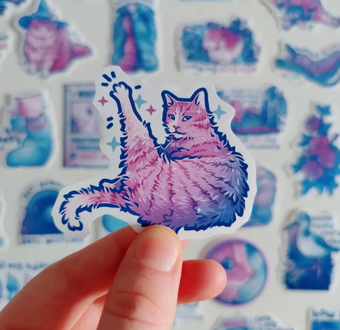 Hand holding up a sticker of a pink tabby cat holding up a leg mid-cleaning