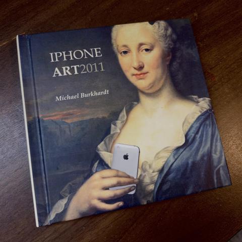 A book titled “iPhone Art 2011” by Michael Burkhardt lies on a wooden surface. The cover features a classical 18th-century portrait of a woman in blue and white period dress, digitally altered to show her holding a white iPhone.