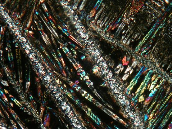 A view of a rock thin section  taken down a polarizing microscope using plane polarized light. The view consists mostly of small, rainbow-coloued feathery, skeletal crystals (clinopyroxene) in a matrix of fine-grained material (former glass), with two larger, long linear crystals of greyer mineral (serpentinized olivine) running NW-SE across central parts of the view.
