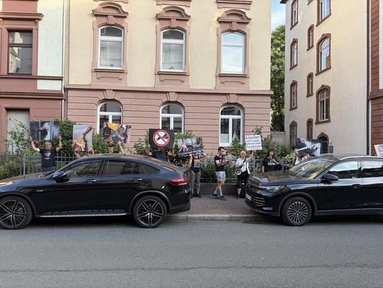 A group of protesters is gathered on a street, holding large signs and banners. Some individuals are using megaphones. They are standing near parked black vehicles and close to a building with large windows. The signs appear to convey messages related to their cause