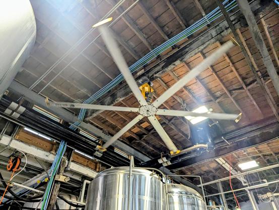 Looking up at a large fan with 6 long blades on a high wooden ceiling with pipes running through . The tops of some large steel tanks are at the bottom of frame