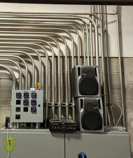 The top of a grey metal cabinet with 2 small grey and black speakers stacked one on top of the other. Behind it on the grey wall is a number of pipes (10+) parallel going up and turning left all in aneat parallel form