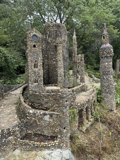 A roughly 7 foot tall structure, which is intended to be a replica of something famous. I’m not sure what though it would be on the little marker at the front of the place.
It’s a building made out of quick Crete, sand and river pebbles. It has small windows and walkways.  Some of the walkways an adult can carefully on down around it

These structures were built by multiple people over a timeframe of about 15 years the man who started it was a sculptor. He was building a small village with his grandchild. It was never meant to be a tourist attraction. It grew serendipitously.