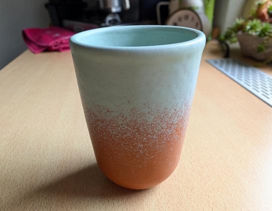 A slightly high-angle, close-up photograph featuring an unglazed ceramic tumbler placed centrally on a light-colored wood grain surface. The tumbler has a textured, orange terracotta base that transitions, via an irregular speckled pattern, to a matte, pale mint-green or light sage-green top rim and interior. The lighting highlights the matte texture of the clay. The background is a gently blurred indoor domestic scene (kitchen counter/desk). Visible in the soft-focus background are a pink cloth, parts of an espresso machine or kitchen appliance, and a terracotta pot with small succulent plants. The overall atmosphere is warm and tactile.
