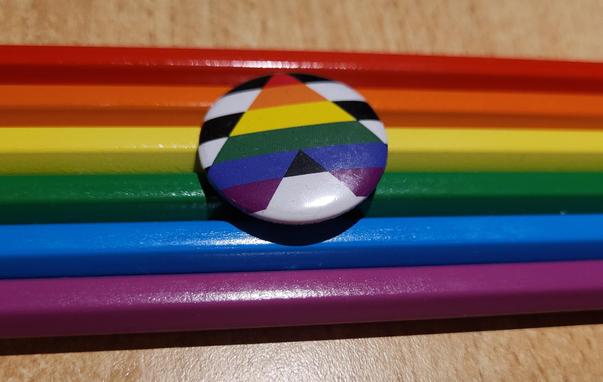 Ein runder Ally-Button mit einem geometrischen Muster liegt auf mehreren horizontal angeordneten Buntstiften. Der Button zeigt verschiedene farbige Segmente in Rot, Orange, Gelb, Grün, Blau, Violett, Schwarz und Weiß, die als Streifen und Dreiecke angeordnet sind. Die Buntstifte liegen unter dem Button in den Farben Rot, Orange, Gelb, Grün, Blau und Violett übereinander übereinander. Das gesamte Arrangement befindet sich auf einer hellen Holzoberfläche.