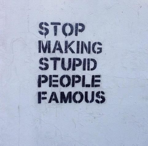 A close-up photograph captures the phrase "STOP MAKING STUPID PEOPLE FAMOUS" in black stencil-style capital letters on a slightly textured. Small cracks and weathering marks are visible across the wall. Minor paint drips are under the first letter. The style suggests street art or graffiti.