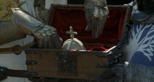 Picture of a small wooden chest with a glimmering round thing with a cross inside, and hands reaching down to pick it up.