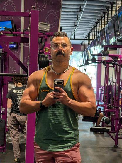 Jorge taking a selfie in front of a gym mirror. His arms are being held close to his upper body which makes them look pretty big and defined. There's when a vein on the left bicep. 

He's wearing a green tank top and pink shorts