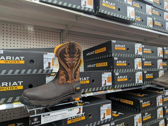 Store shelves with black shoe boxes with Ariat brand "Work" boots and one boot on display with fancy embroidered ankle and upward top part