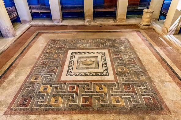 Roman mosaic floor featuring a central square panel depicting a small vessel with two birds perched on its rim. The panel is surrounded by a decorative border of interlocking geometric patterns in black, white, red, and yellow. The mosaic is set within a site with stone columns, evoking a preserved archaeological setting.