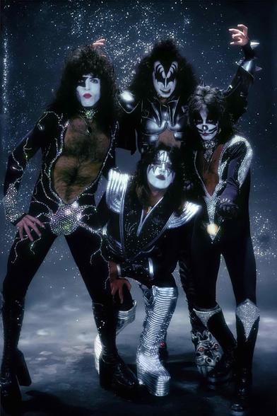 vintage promo photo of the band KISS, they are all wearing black and silver spandex with rhinestones, platform boots, long styled hair, and their trademark intricate makeup. they seem to be on a set with fog machines going and all kinds of twinkly shit behind them. they…look pretty fem for a bunch of macho dudes.