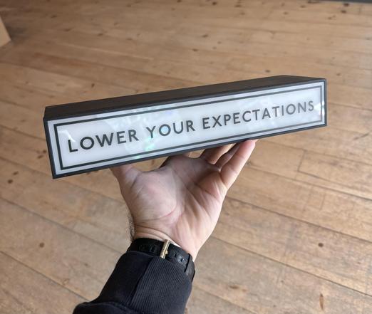Photo of a backlit light sign that reads “Lower Your Expectations” being held in someone’s hand against a wooden floor