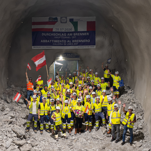 A large group of tunnel construction workers in high-visibility vests and hard hats celebrate inside a newly broken-through tunnel, waving Austrian and European Union flags beneath a commemorative banner.