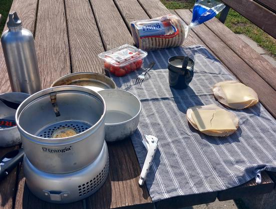Ett typiskt campingbord med ett trangiakök, och på en kökshandduk står en mugg och ett par mackor
