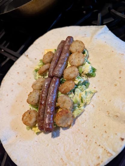 4 sausage links and tater tot coins rest atop scrambled eggs and cheese on a tortilla