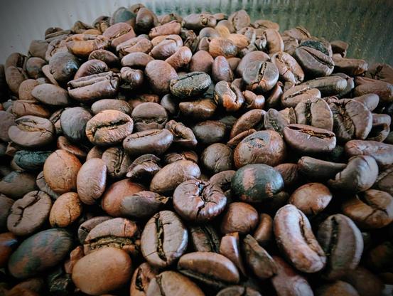 Eine Nahaufnahme aus der Vogelperspektive zeigt einen Haufen gerösteter Kaffeebohnen, die den gesamten Bildausschnitt füllen. Die Bohnen weisen unterschiedliche Röstgrade auf – von hellbraun bis dunkelbraun – mit sichtbaren Einkerbungen und teils leicht seidigem Glanz. Das Licht fällt zentral auf die Bohnen, während die Ränder des Bildes leicht abgedunkelt sind.
