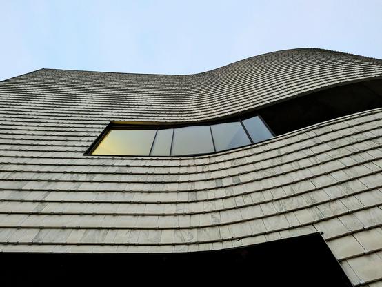 Looking up from close, almost under, at a tall building that is slightly curved. Most of the building facade is covered with an overlapping off white wood siding. A glass window well above and with siding above and below is in the middle of the building viewed above - the glass is reflecting a slight yellow of the late evening sky, with the gently curving portion to the right of the window - dark and likely a balcony space.