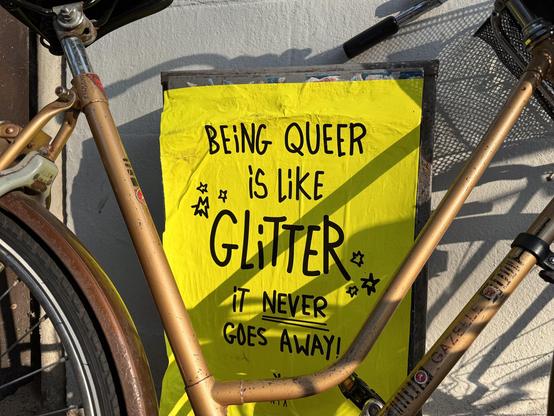 bright yellow poster, slightly obscured by a bronze bicycle, reads in black lettering “being queer is like glitter, it never goes away!”