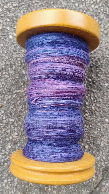 A spinning wheel bobbin about half filled with fine singles in shades of blueish purples. There's a light shine to the yarn.