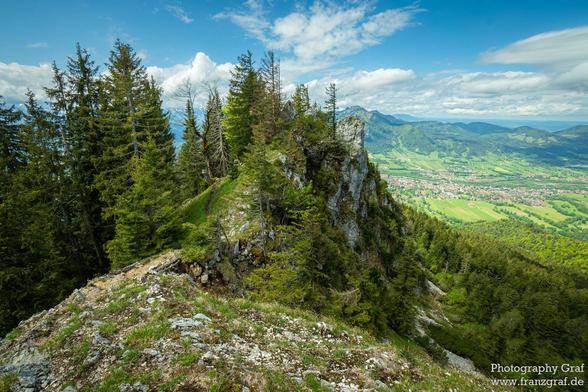This image captures a breathtaking and expansive mountain landscape, showcasing the serene beauty of a high-altitude vista. The foreground features a rugged, rocky cliff adorned with patches of green vegetation, including moss and small shrubs, adding texture and a sense of wildness to the scene.
Beyond the cliff, the landscape opens up to reveal a vast expanse of dense forests and rolling hills. The forests are dominated by tall evergreen trees, their dark green foliage creating a rich and vibrant tapestry. The hills stretch out into the distance, their gentle slopes covered in a mix of green meadows and patches of woodland.
In the far background, a valley unfolds with a mix of green fields, clusters of buildings, and winding roads, suggesting a rural or semi-urban settlement nestled within the natural landscape. The buildings are scattered across the valley, their red and brown roofs contrasting with the surrounding greenery.
The sky above is a brilliant blue, dotted with fluffy white clouds that add depth and a sense of openness to the scene. The sunlight bathes the entire landscape in a bright, natural light, highlighting the vibrant colors of the forests, the rocky cliff, and the distant valley. The interplay of light and shadow adds texture and emphasizes the dramatic beauty of the landscape.