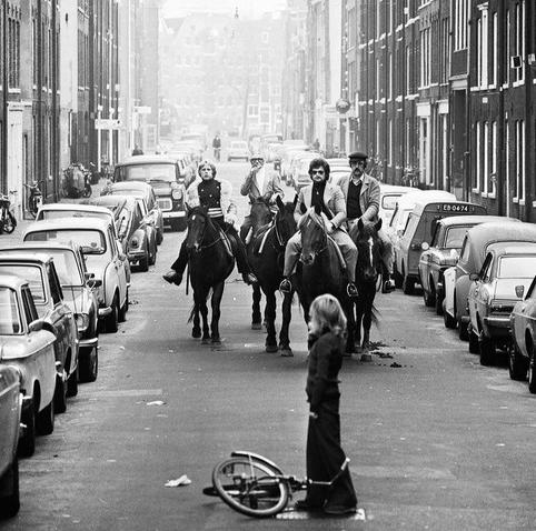 A black and white photo of an Amsterdam Street with cars parked at both sides. 4 horsemen in time typical urban attire riding slowly in the middle of the street towards the viewer, in the foreground, still about 15 meters in front of them, a blond child is standing, looking at the riders.
