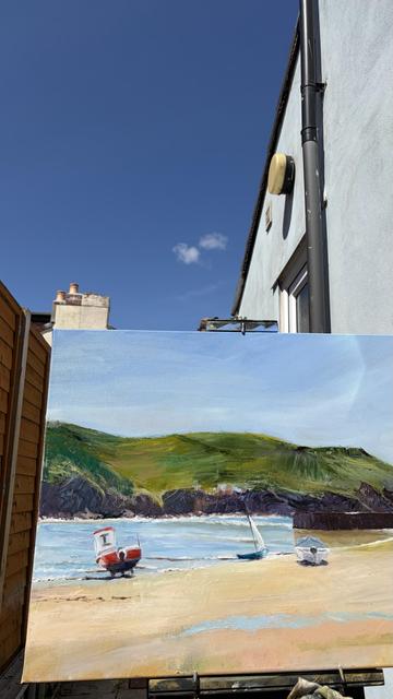 A vibrant painting of a beach scene featuring two boats on the sandy shore. In the background, there are rolling green hills under a bright blue sky with a few clouds. A building can be seen at the side, showing that the artist is actually painting in their garden