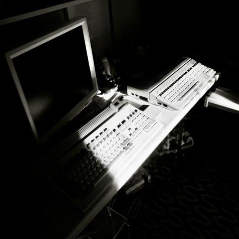 A black‑and‑white photo of a retro computer setup on a desk. Two vintage machines—a Commodore Amiga and an Atari ST—sit side by side, each with integrated keyboards and distinctive ridged casings. A blank monitor stands to the left, partly illuminated by a diagonal beam of sunlight cutting across the desk. Small figurines rest near the monitor, and a patterned carpet is visible below, adding texture to the nostalgic scene.
