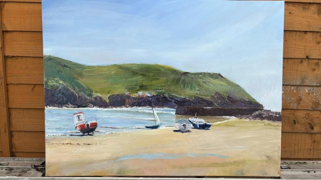 A seaside landscape painting featuring a calm beach with several boats on the sand. Green hills rise in the background under a clear blue sky. The artwork is balanced against a wooden garden fence.