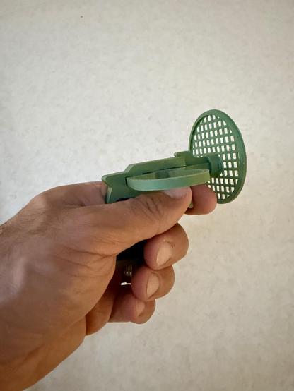 An immaculate left Arab hand holds a small green 3D‑printed fly swatter. The swatter has a short handle and a circular, grid‑patterned striking surface.